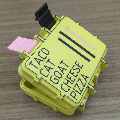 Oblique angle of a rugged-looking hinged yellow box, approximately the size and shape required to hold a deck of cards. It has two hinges, one pink and one black. On the front in landscape, in black text, it says "Taco Cat Goat Cheese Pizza". To the right of that are three vertical lines, the first two black and the third pink. The middle line is slightly longer.