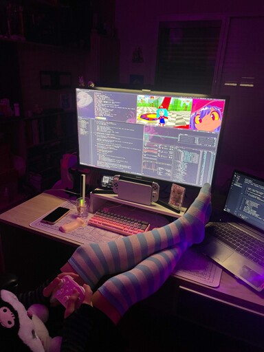 Person holding a Kuromi plush and a pink controller, wearing striped thigh high socks and resting their legs on a desk with a large monitor and a laptop running Gentoo Linux with bspwm. On screen there is code, btop, fastfetch, Niko from Oneshot and multiplayer SM64 with a Hatsune Miku skin.